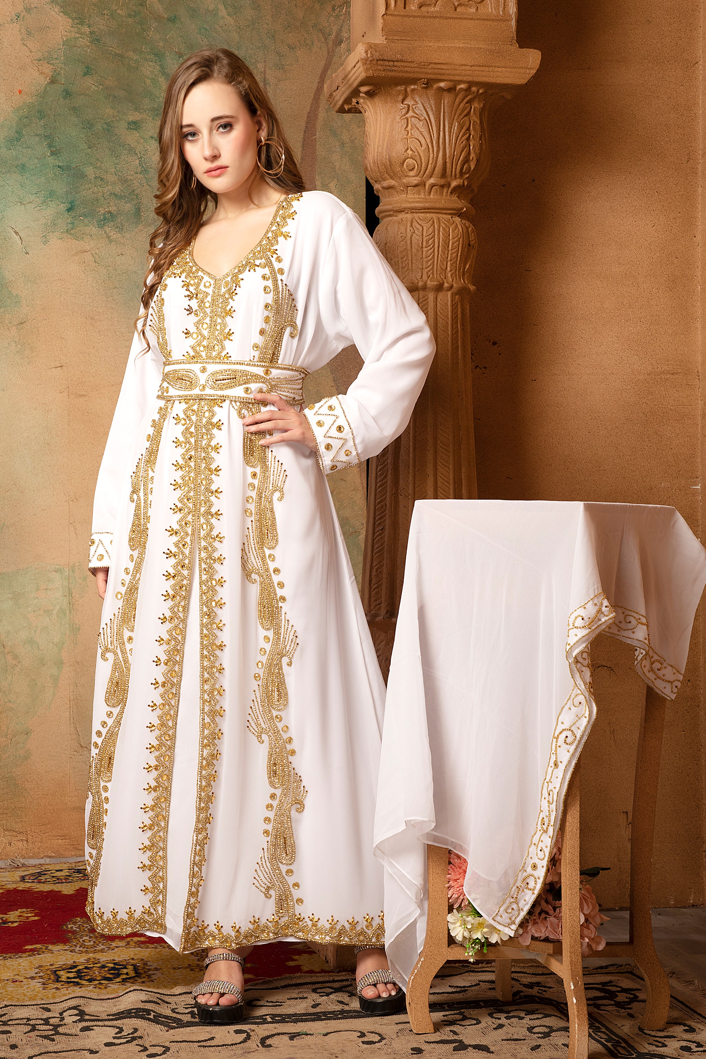 A woman standing in a traditional, embellished Moroccan caftan with golden beads, posing next to a white caftan fabric displayed on a chair.