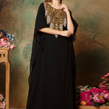 A woman standing in a traditional black kaftan with embroidered details and a scarf, posing against a floral backdrop.