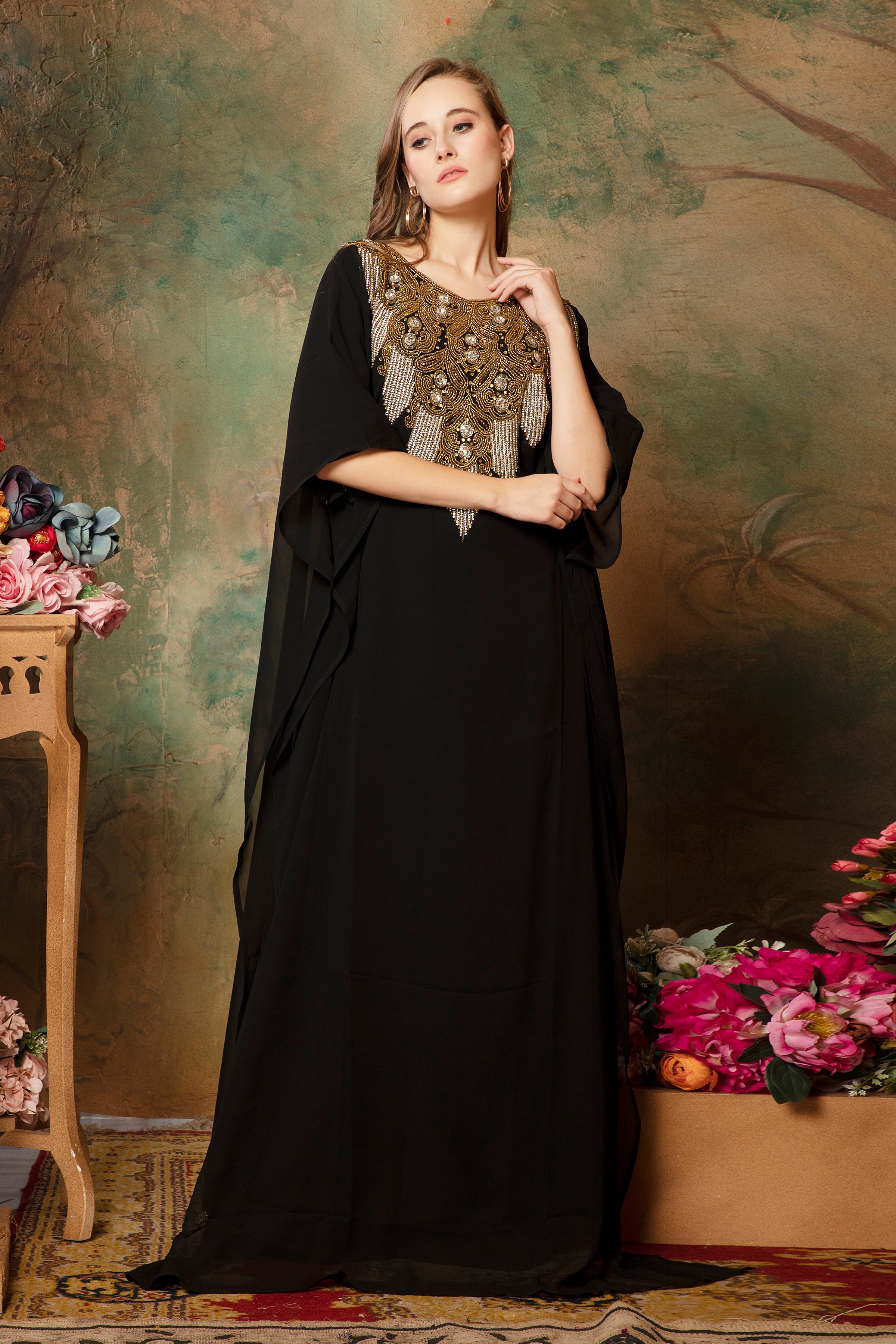A woman standing in a traditional black kaftan with embroidered details and a scarf, posing against a floral backdrop.