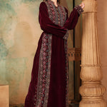 A woman standing next to a column, wearing a traditional, embroidered, dark purple gown with long sleeves and a straight neckline. The gown features geometric patterns and is adorned with beads.