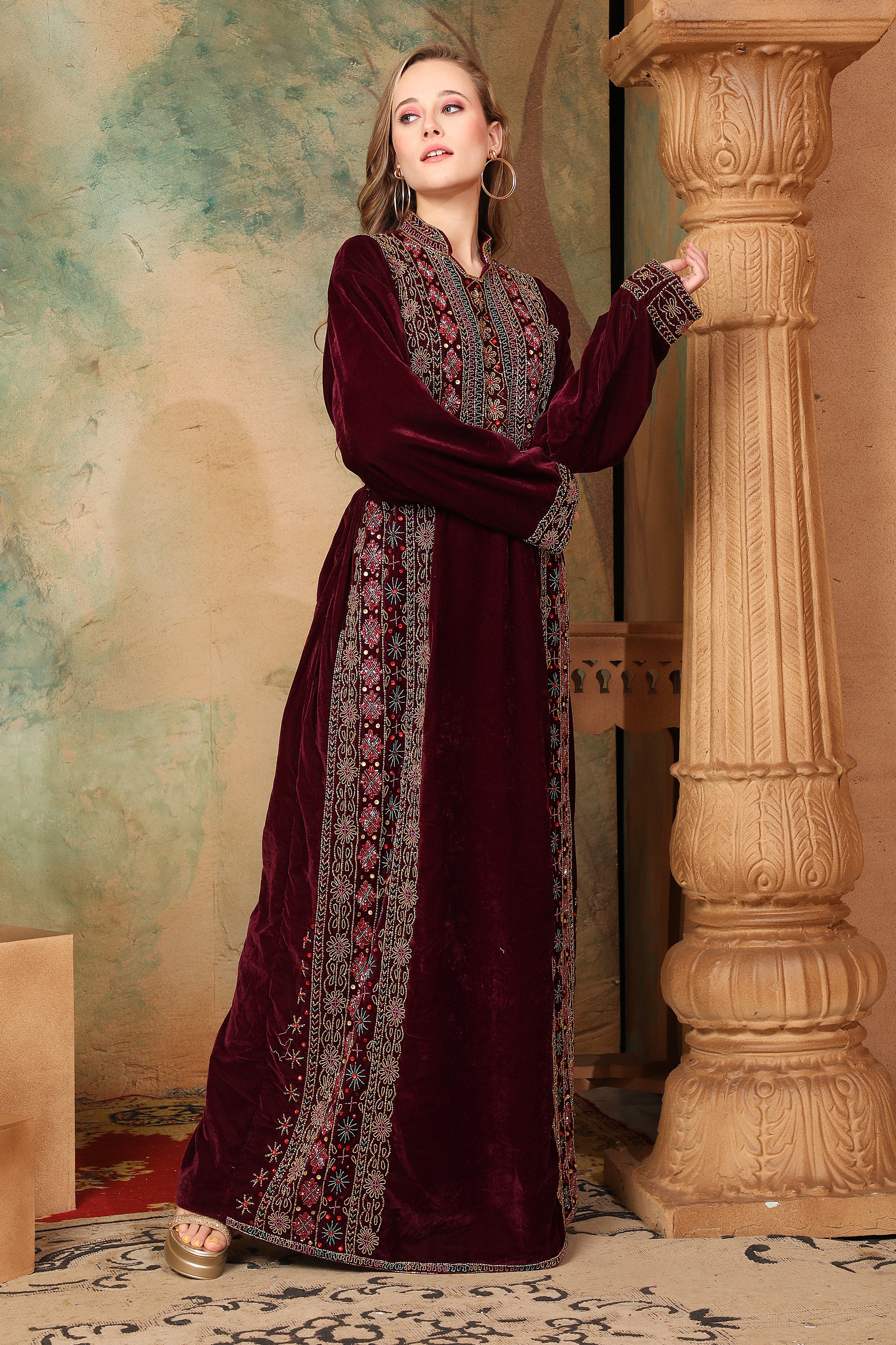 A woman standing next to a column, wearing a traditional, embroidered, dark purple gown with long sleeves and a straight neckline. The gown features geometric patterns and is adorned with beads.