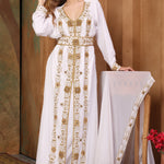 A woman wearing a traditional Moroccan white caftan with golden bead embellishments, posing in a room with cultural decor.