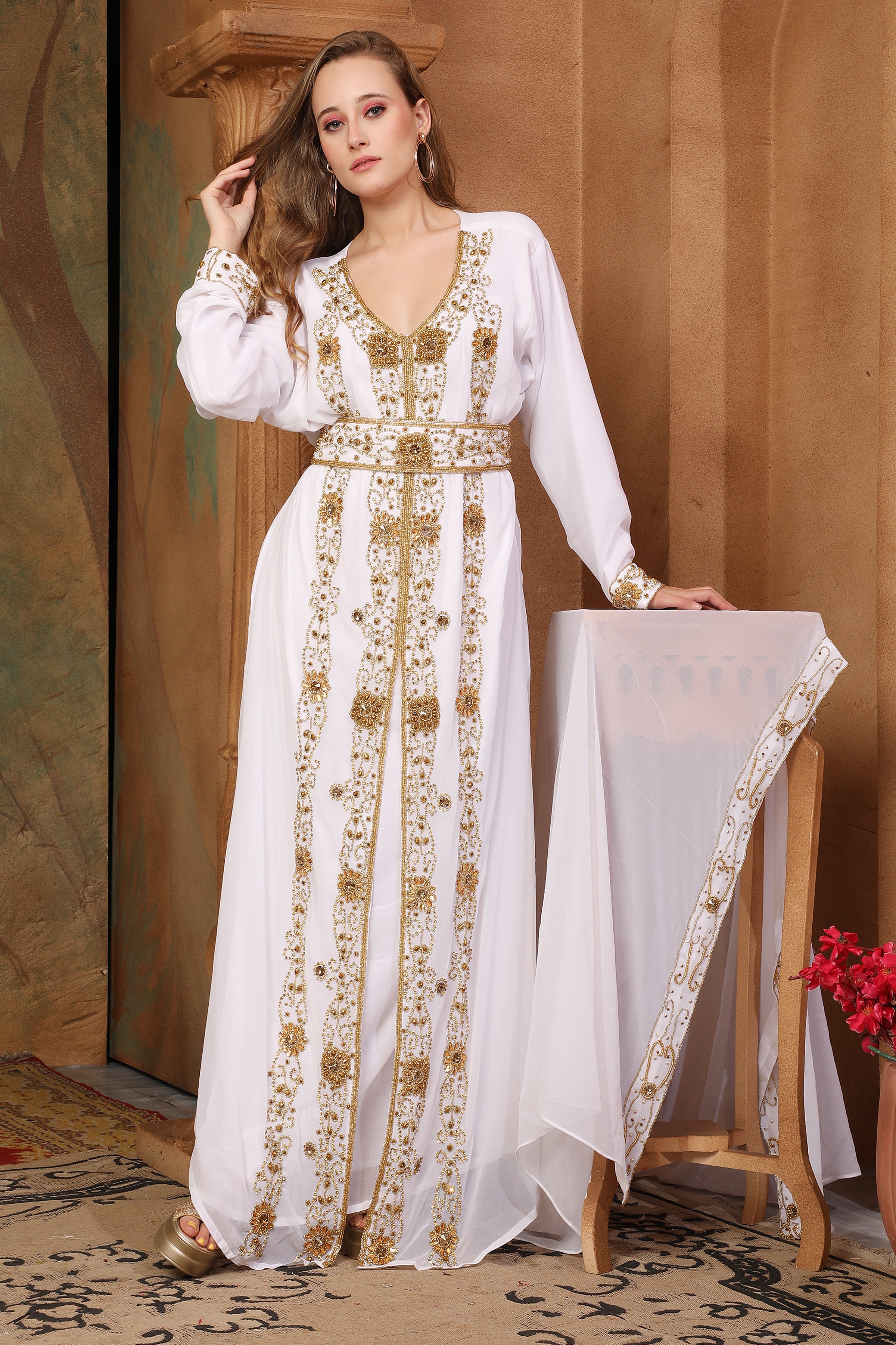 A woman wearing a traditional Moroccan white caftan with golden bead embellishments, posing in a room with cultural decor.