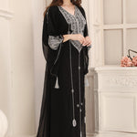 A woman wearing a black Moroccan caftan with silver embellishments, standing in a room with a white fireplace.