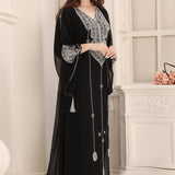 A woman wearing a black Moroccan caftan with silver embellishments, standing in a room with a white fireplace.