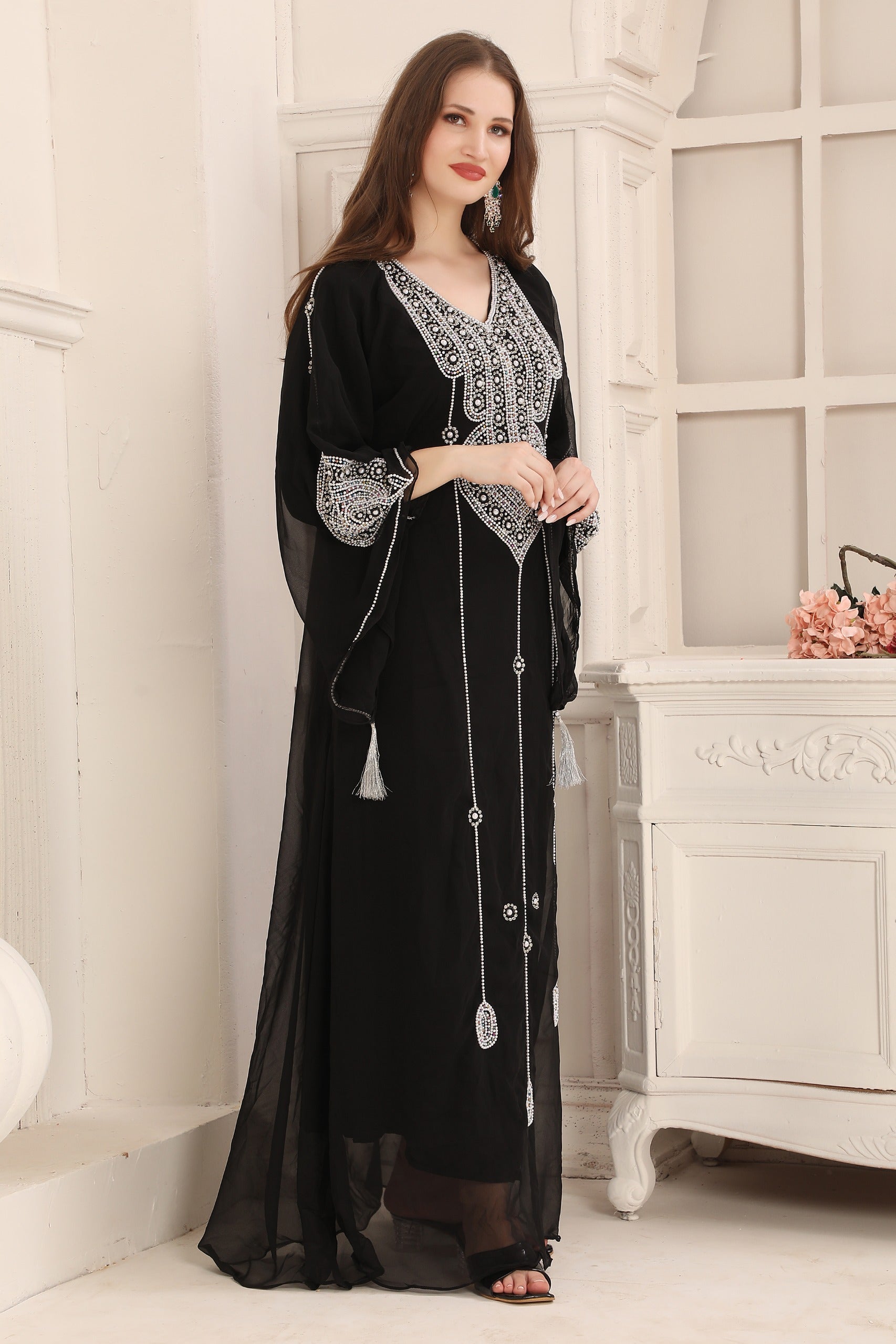 A woman wearing a black Moroccan caftan with silver embellishments, standing in a room with a white fireplace.