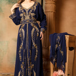 A woman wearing a navy blue, embellished with golden beads, long-sleeve Moroccan caftan with a belt, standing next to a floral arrangement.