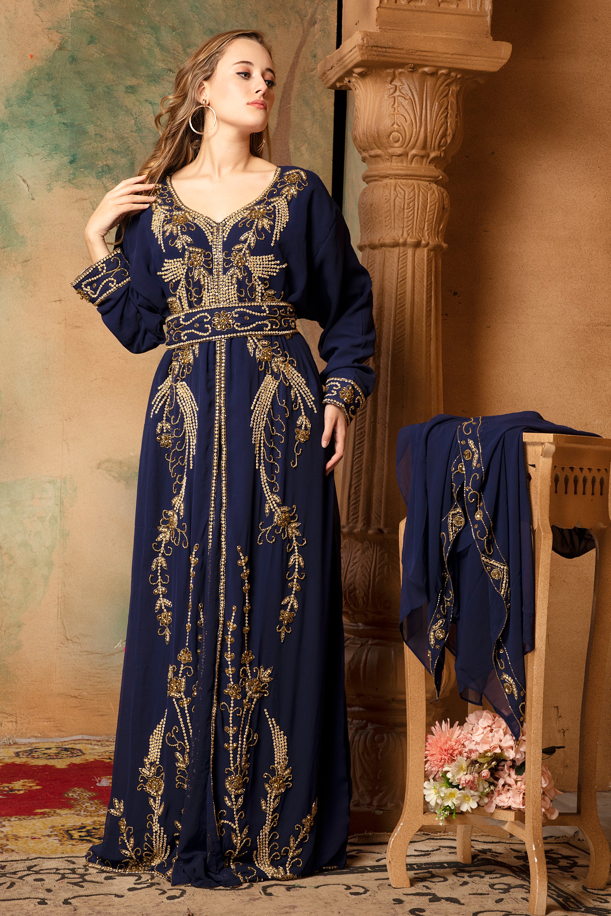 A woman wearing a navy blue, embellished with golden beads, long-sleeve Moroccan caftan with a belt, standing next to a floral arrangement.