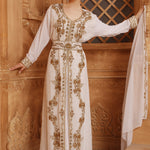 A woman wearing a traditional Moroccan caftan with gold color beads and embroidery, standing in front of wooden panel background.