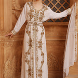 A woman wearing a traditional Moroccan caftan with gold color beads and embroidery, standing in front of wooden panel background.