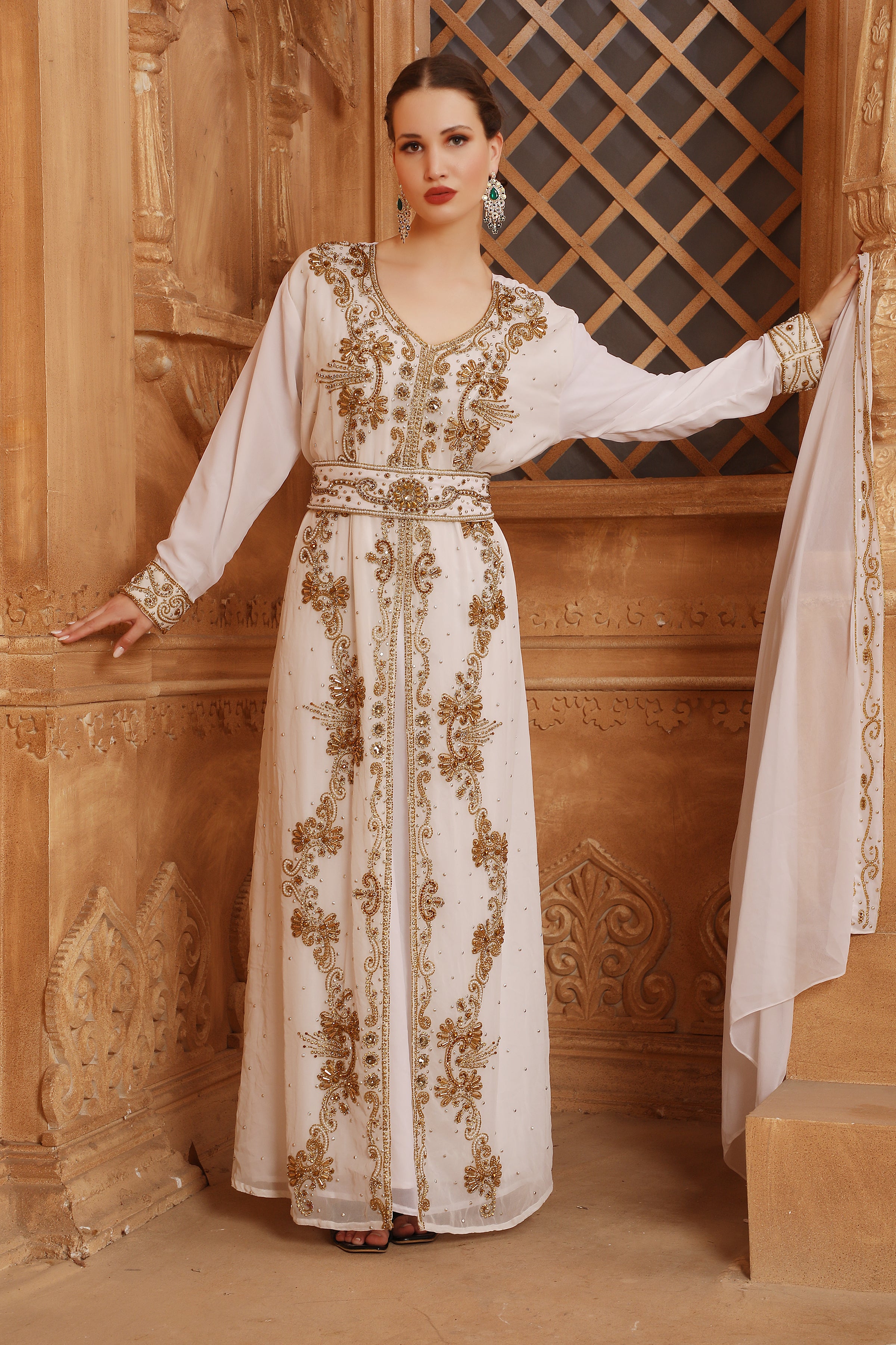 A woman wearing a traditional Moroccan caftan with gold color beads and embroidery, standing in front of wooden panel background.