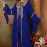 A woman standing in a beautiful blue caftan with golden bead embellishments, posing in a traditional, elegant style.