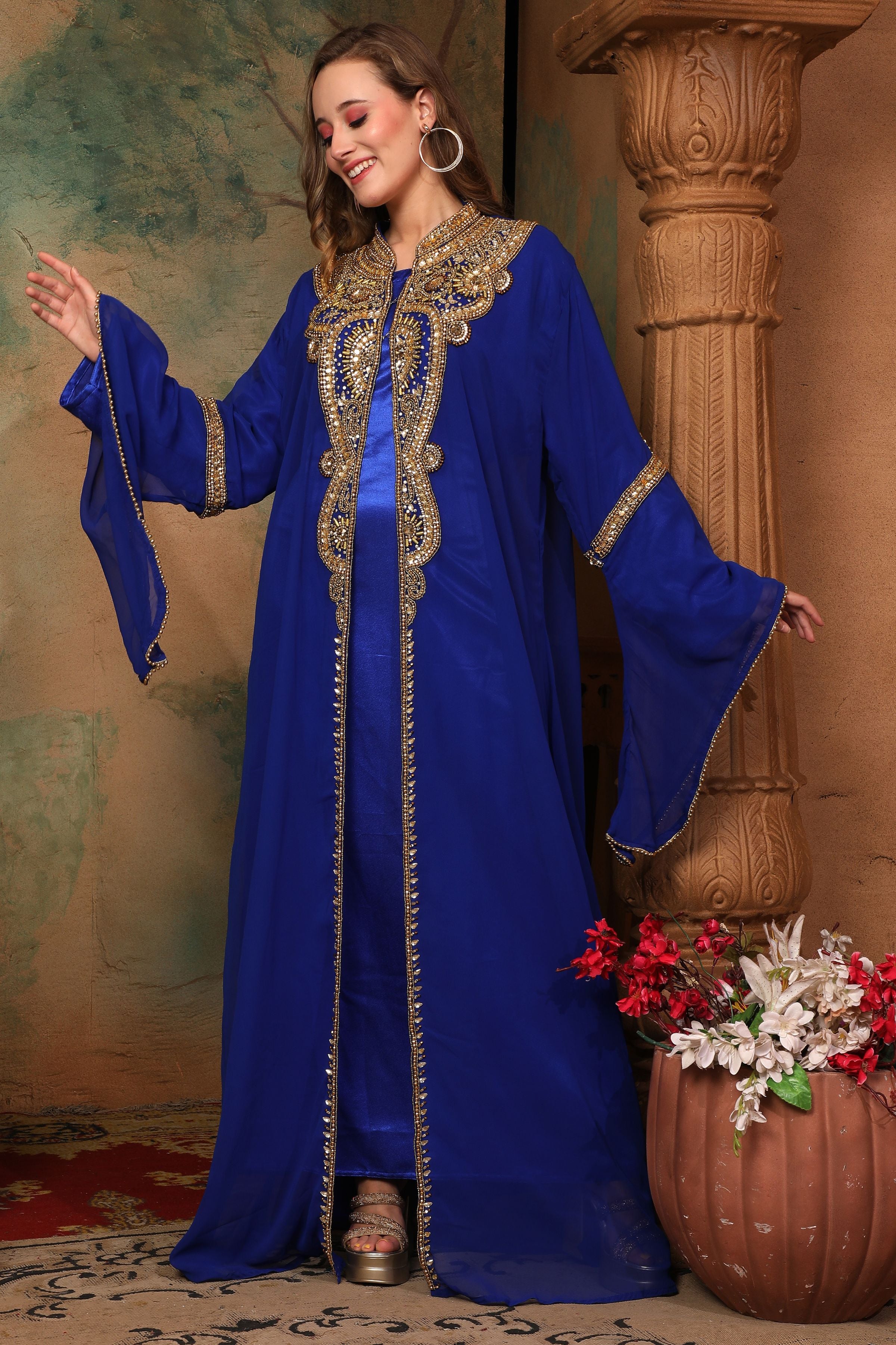 A woman standing in a beautiful blue caftan with golden bead embellishments, posing in a traditional, elegant style.