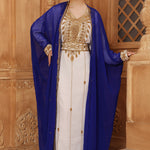 A woman wearing a traditional Moroccan caftan with gold color beads, in a white and blue color scheme, standing in a room with wooden panel walls.