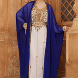 A woman wearing a traditional Moroccan caftan with gold color beads, in a white and blue color scheme, standing in a room with wooden panel walls.