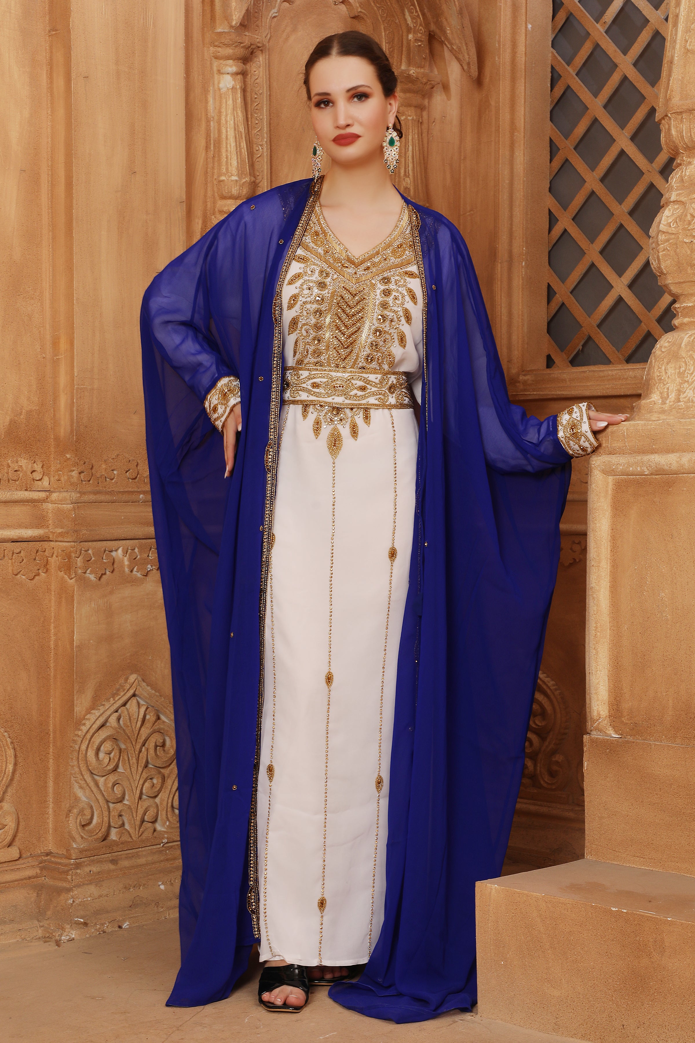 A woman wearing a traditional Moroccan caftan with gold color beads, in a white and blue color scheme, standing in a room with wooden panel walls.