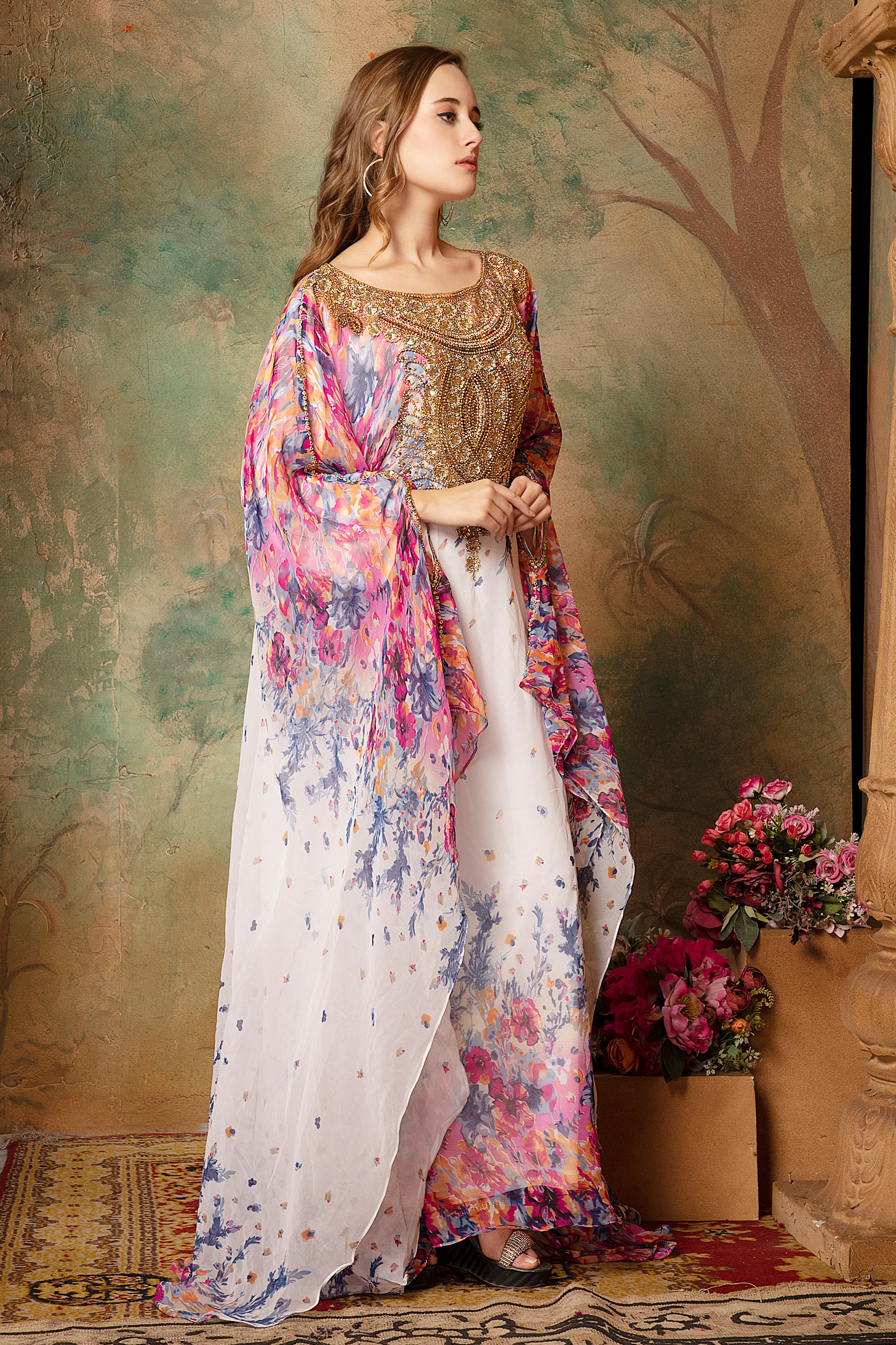 A woman wearing a multicolored floral printed kaftan dress with long sleeves, posing against a green floral backdrop with a tree in the background. The dress features geometric patterns and is embellished with golden beads.