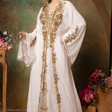 A woman wearing a traditional Moroccan white caftan with golden bead embellishments, posing against a green floral backdrop.
