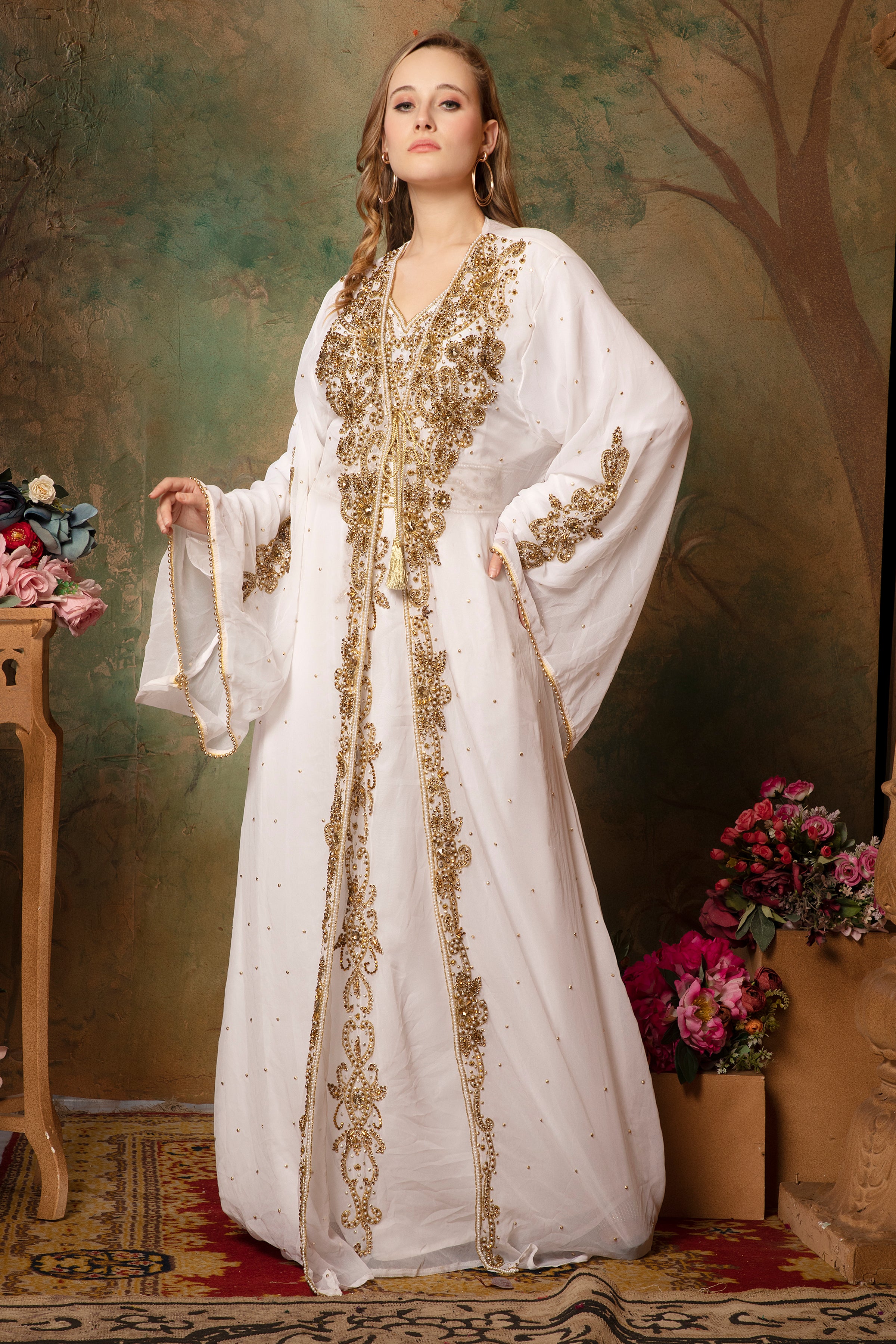 A woman wearing a traditional Moroccan white caftan with golden bead embellishments, posing against a green floral backdrop.