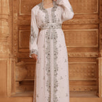 A woman standing in a room with wooden walls wearing a traditional Moroccan silver-beaded caftan with long sleeves.