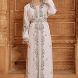 A woman standing in a room with wooden walls wearing a traditional Moroccan silver-beaded caftan with long sleeves.