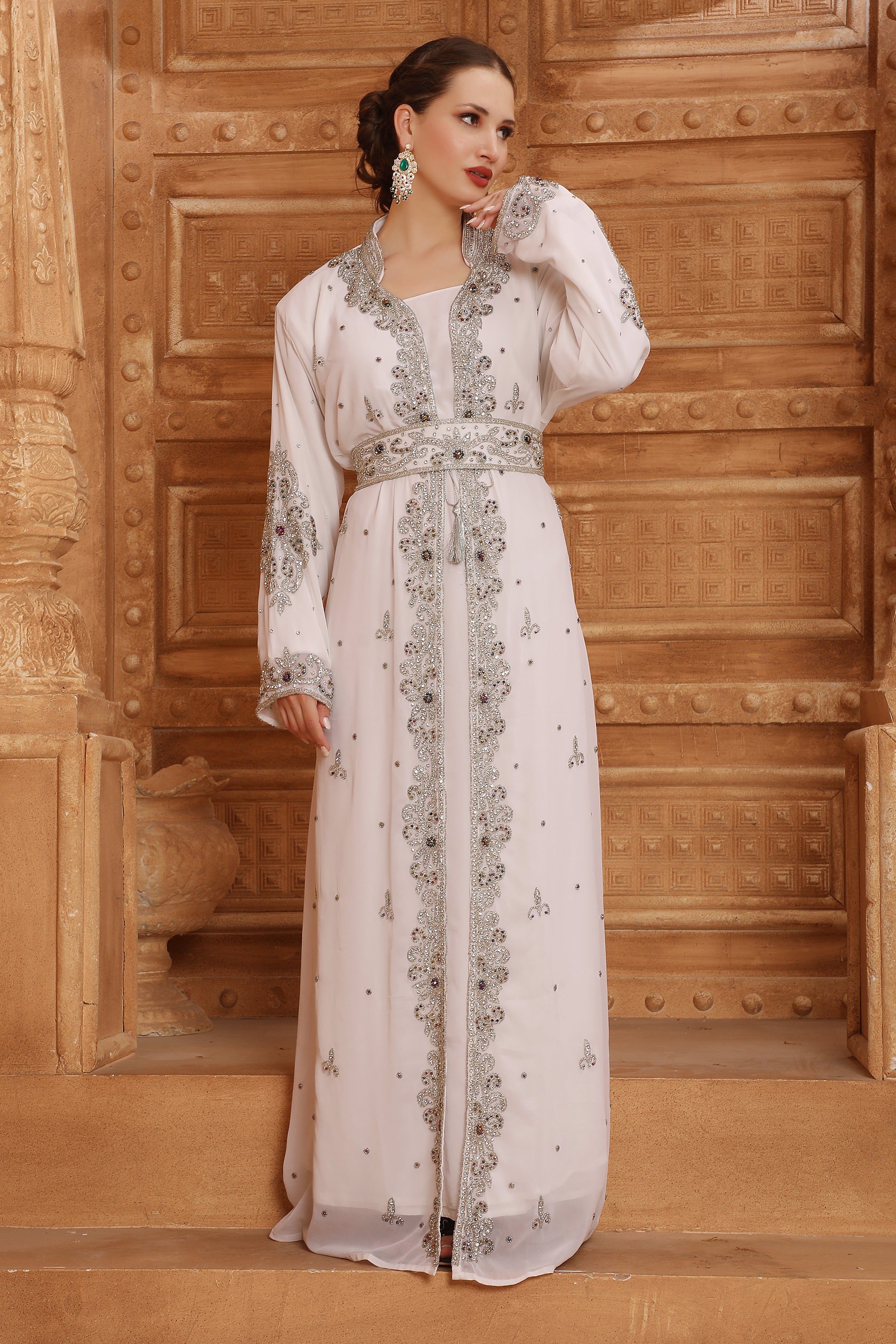A woman standing in a room with wooden walls wearing a traditional Moroccan silver-beaded caftan with long sleeves.