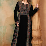 A woman wearing a traditional, embroidered black gown with decorative beadwork, posing next to a column in a room with a painted wall.