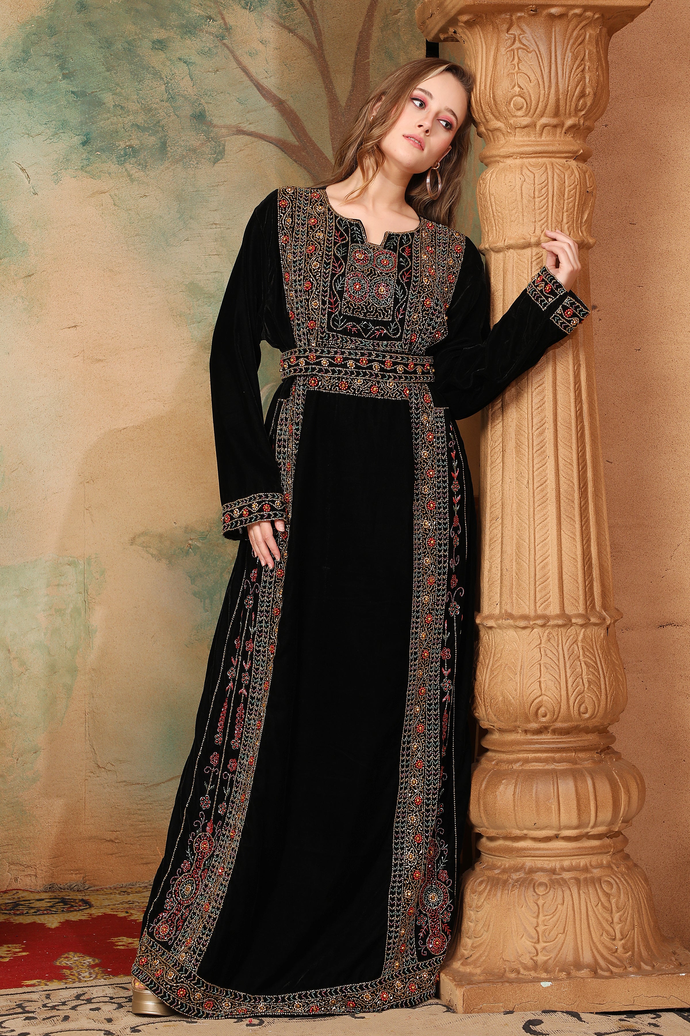 A woman wearing a traditional, embroidered black gown with decorative beadwork, posing next to a column in a room with a painted wall.