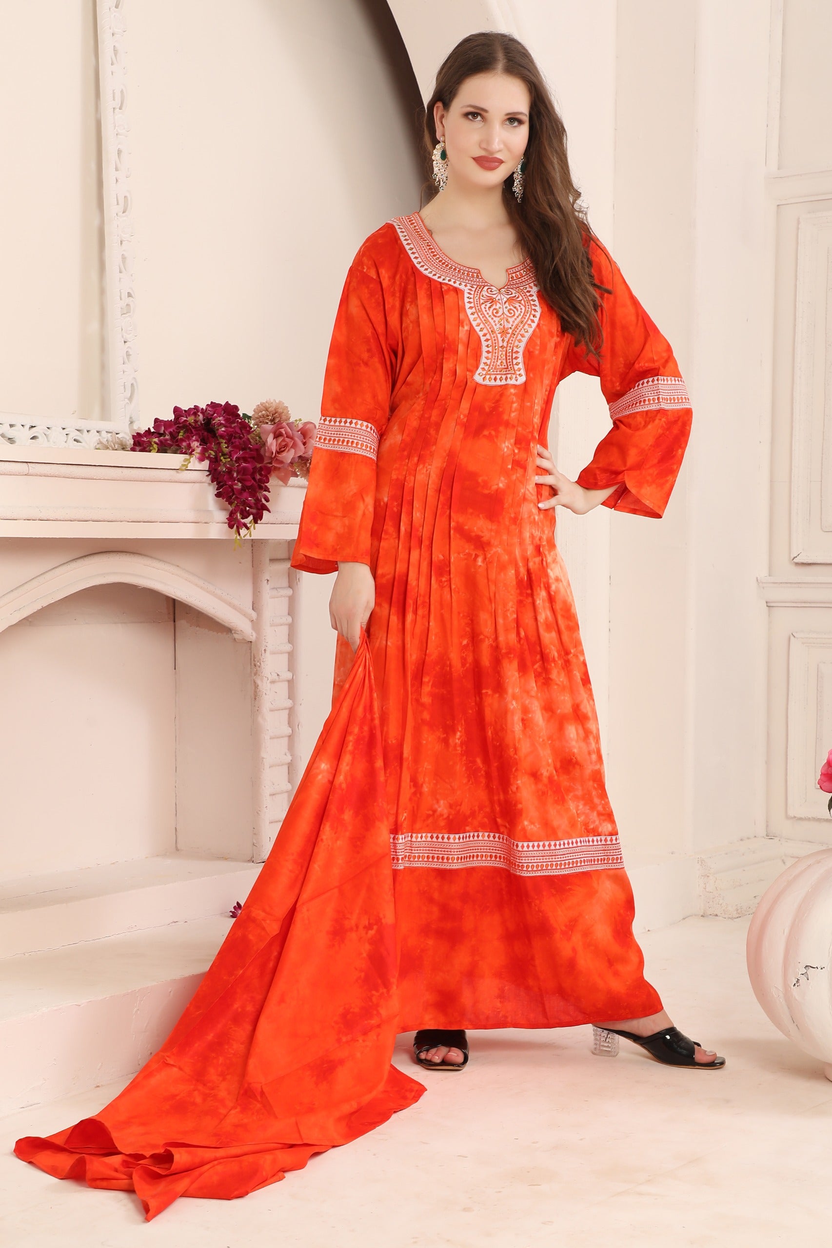A woman modeling a red cotton kaftan with Aari work with beads, standing in a room with a white fireplace.
