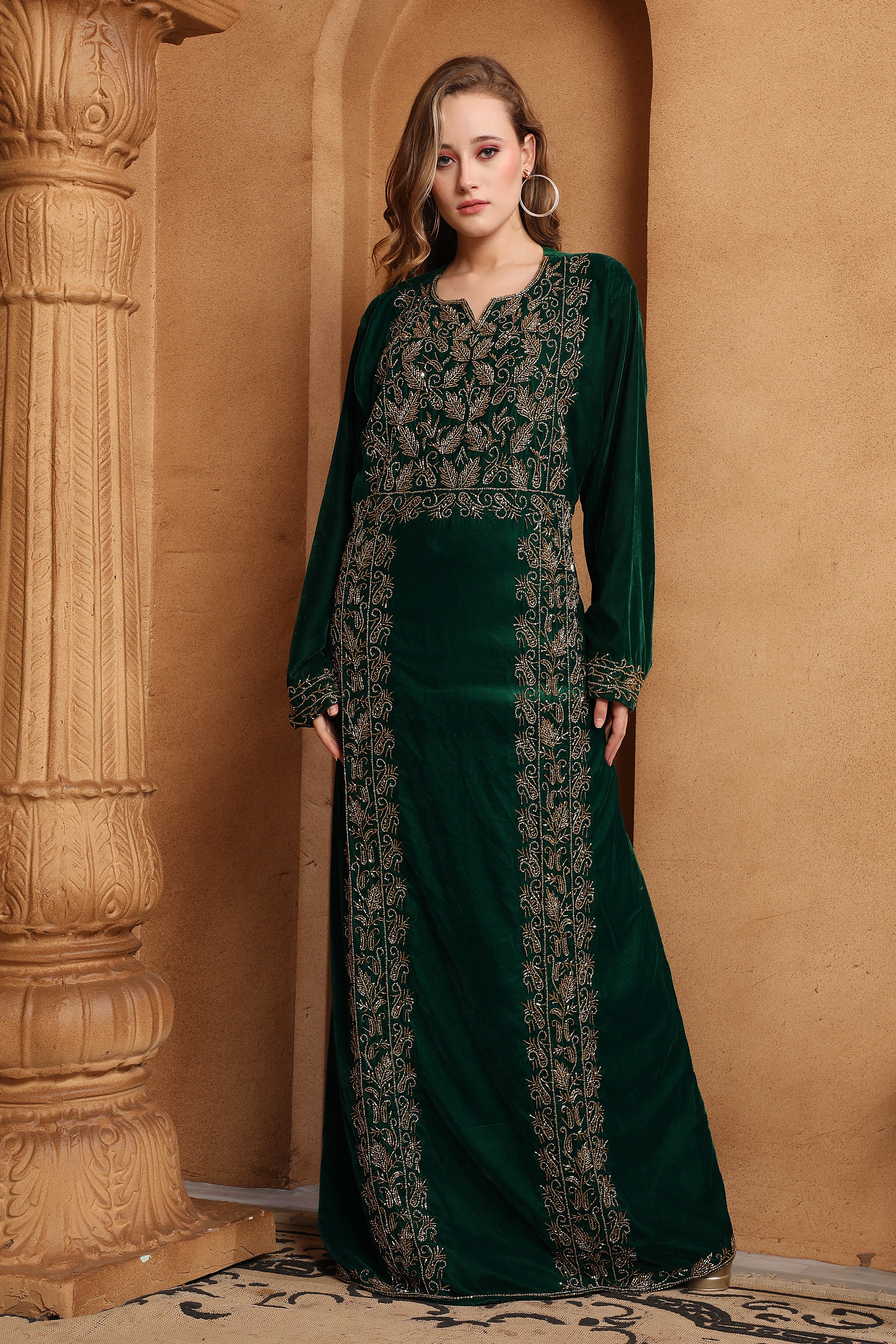 A woman wearing a green embroidered velvet gown with long sleeves, featuring geometric and floral patterns, posing in a columned room.