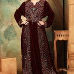 A woman standing in a beautifully embroidered brown velvet gown with geometric patterns, featuring long straight sleeves and a belt.