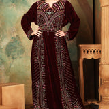 A woman standing in a beautifully embroidered brown velvet gown with geometric patterns, featuring long straight sleeves and a belt.