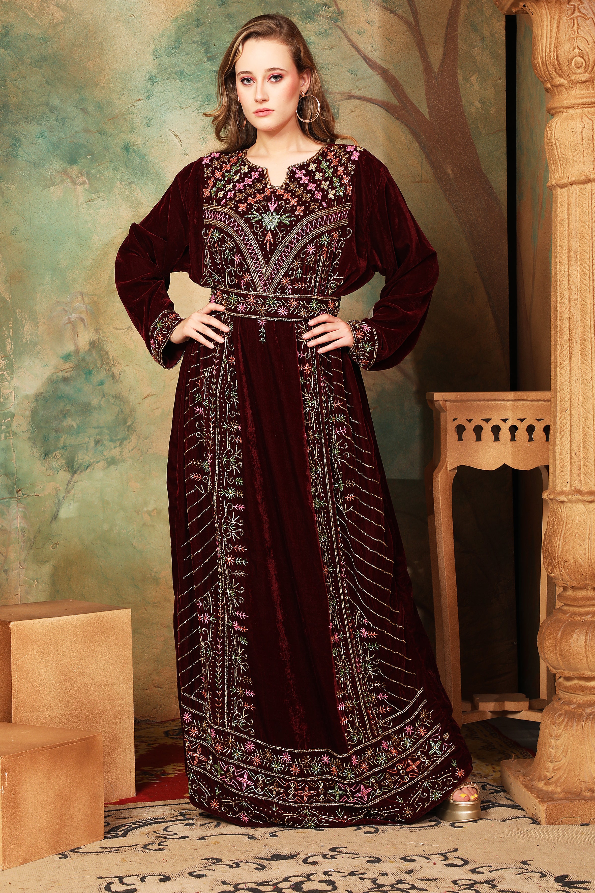 A woman standing in a beautifully embroidered brown velvet gown with geometric patterns, featuring long straight sleeves and a belt.