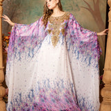 A woman standing in a beautifully printed kaftan dress with long straight sleeves and decorative golden beads, posing in a room with a floral patterned backdrop.