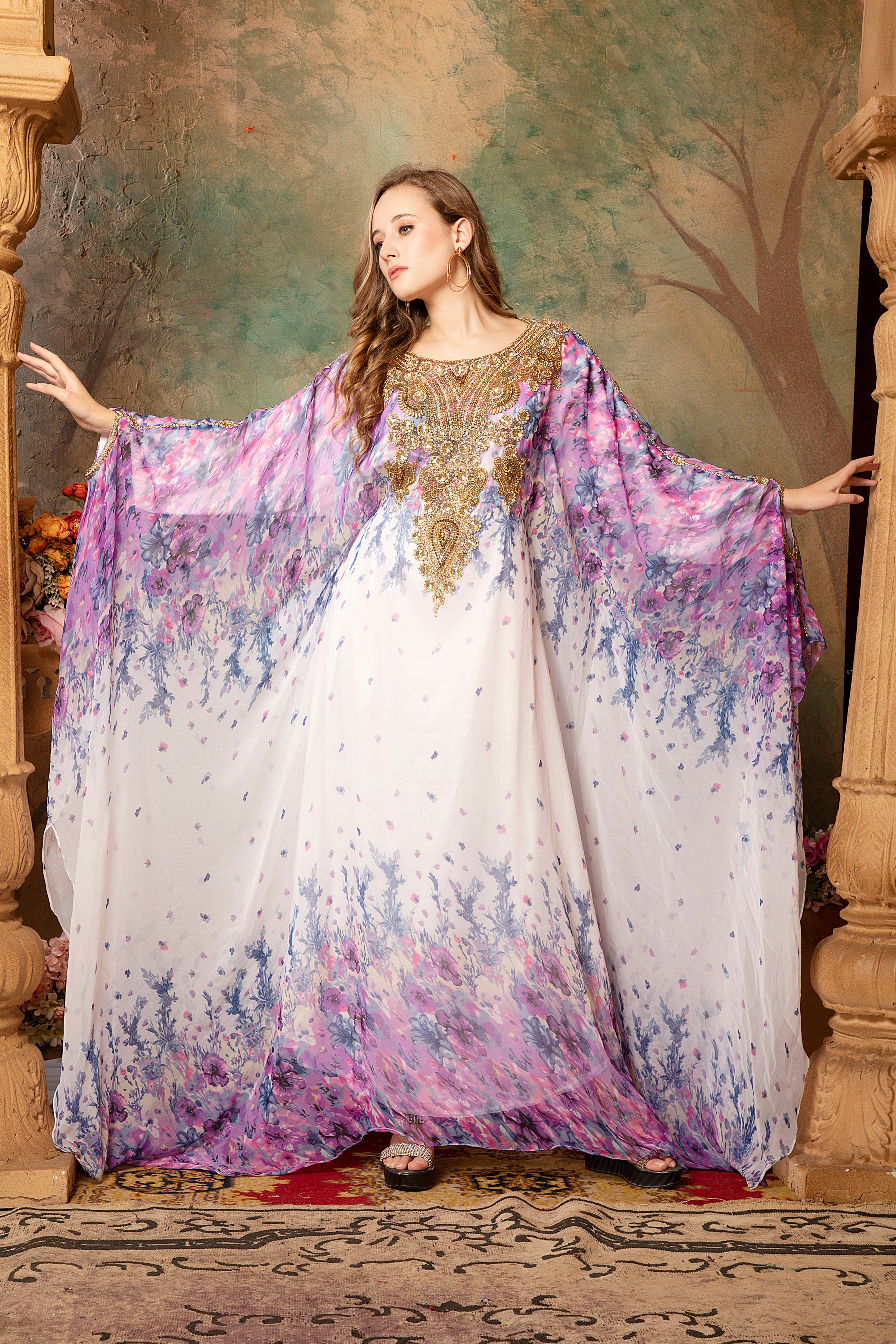 A woman standing in a beautifully printed kaftan dress with long straight sleeves and decorative golden beads, posing in a room with a floral patterned backdrop.