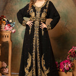 A woman standing in a traditional, embellished Moroccan caftan with black fabric and gold geometric patterns, posing against a floral backdrop.