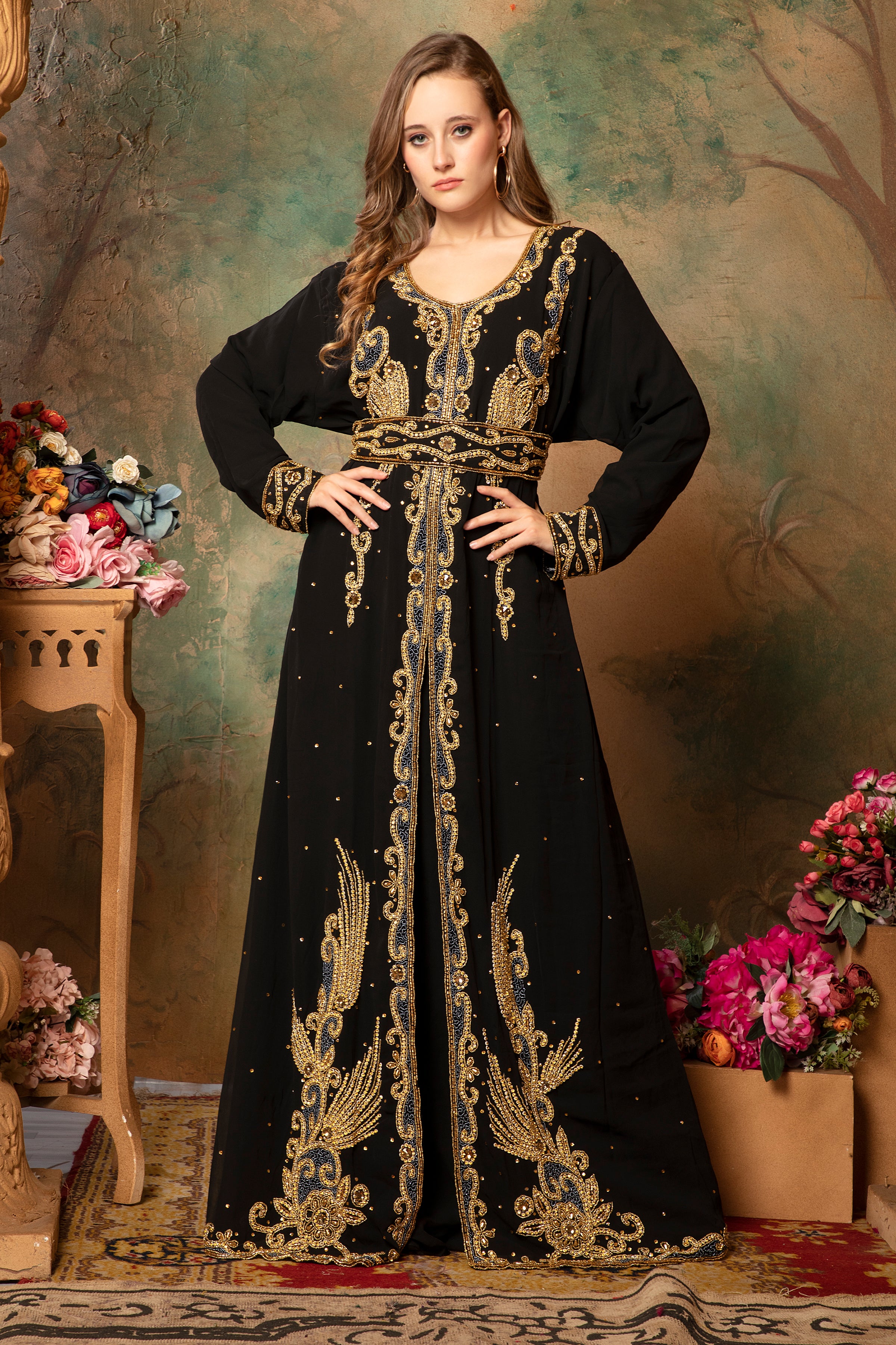 A woman standing in a traditional, embellished Moroccan caftan with black fabric and gold geometric patterns, posing against a floral backdrop.