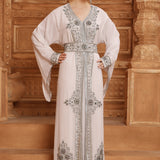 A woman standing in front of a wooden backdrop wearing a white and silver Moroccan caftan with long sleeves and intricate beadwork.
