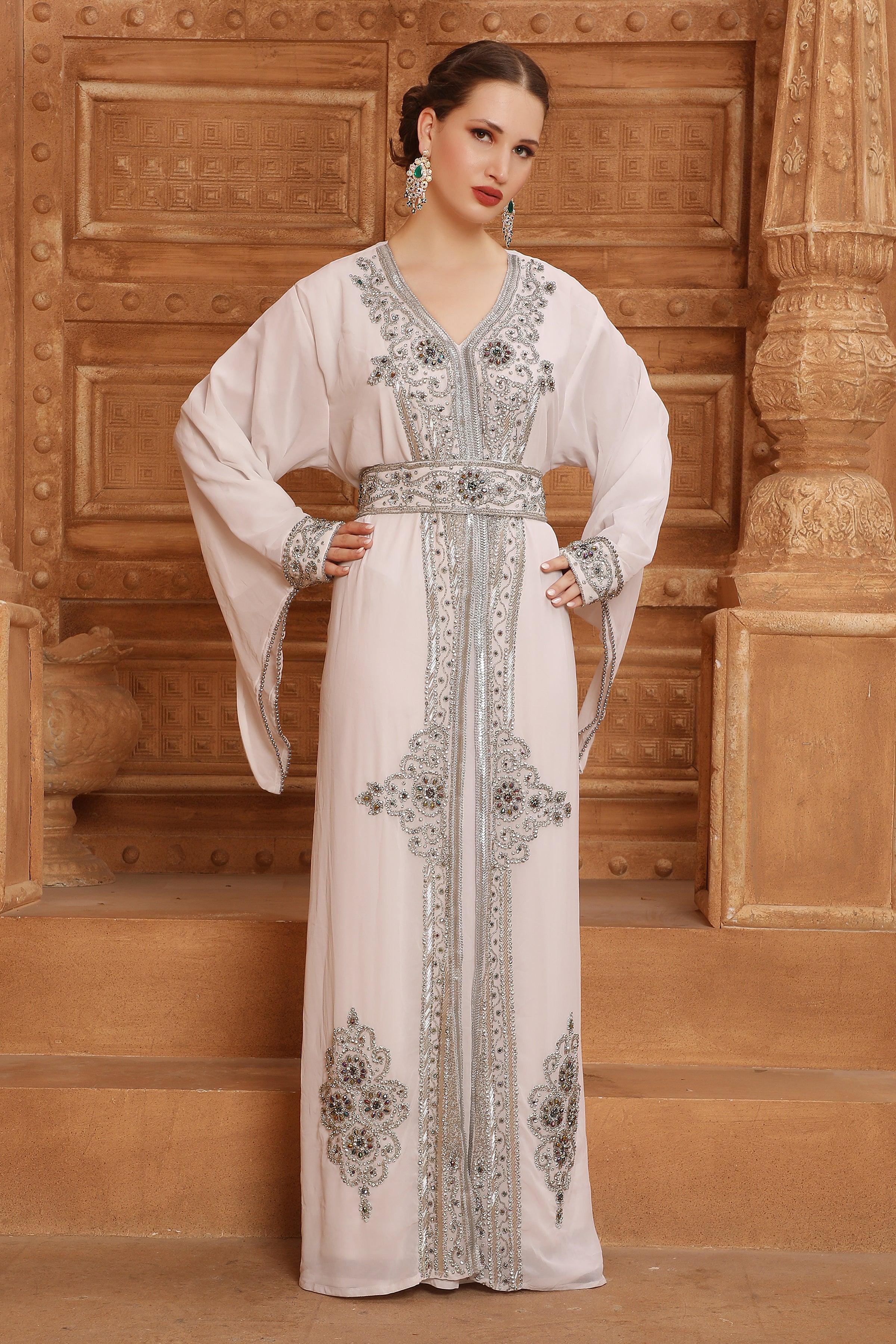 A woman standing in front of a wooden backdrop wearing a white and silver Moroccan caftan with long sleeves and intricate beadwork.