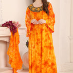 A woman standing in a room wearing a yellow and orange floral kaftan with gold bead detailing around the neckline.