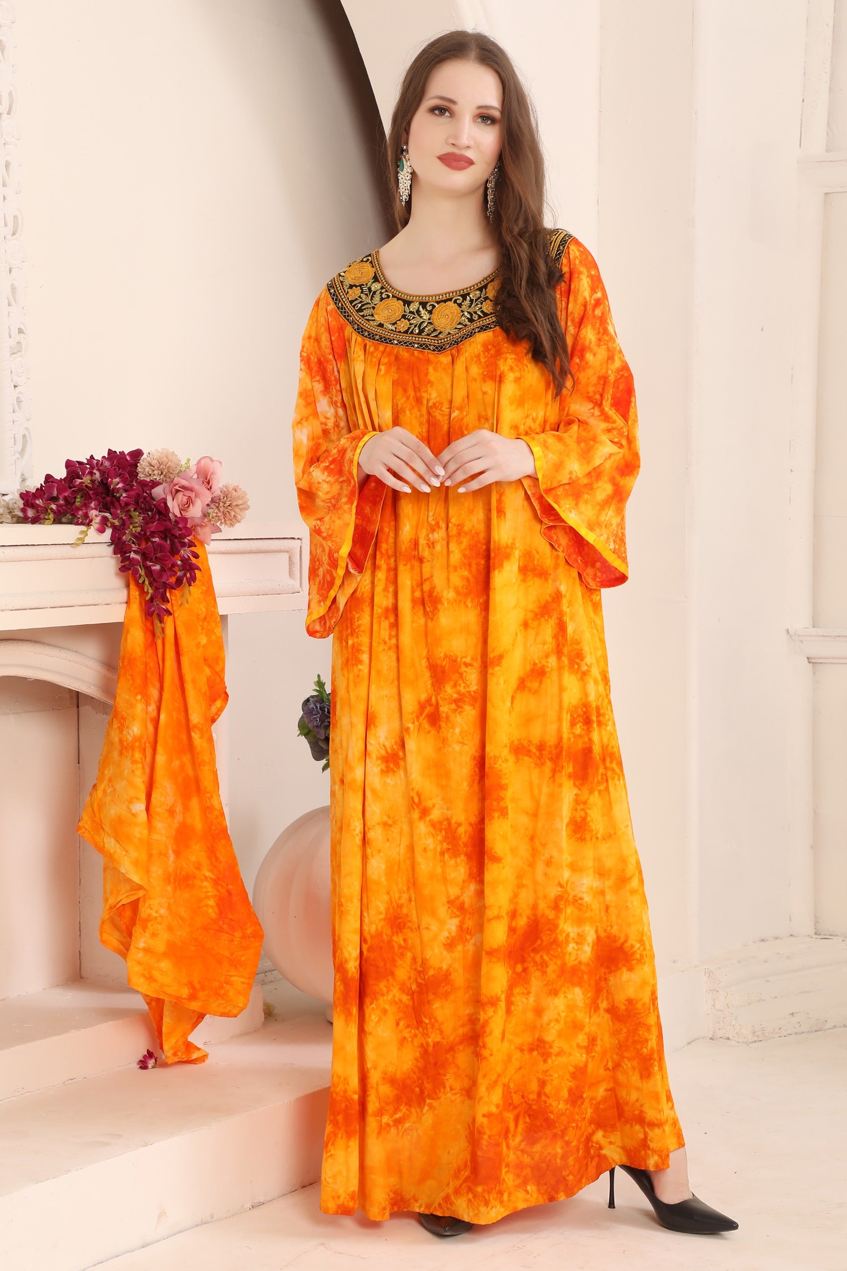 A woman standing in a room wearing a yellow and orange floral kaftan with gold bead detailing around the neckline.