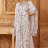 A woman wearing a white and silver Moroccan caftan with floral patterns and long sleeves, standing in a room with wooden decor.