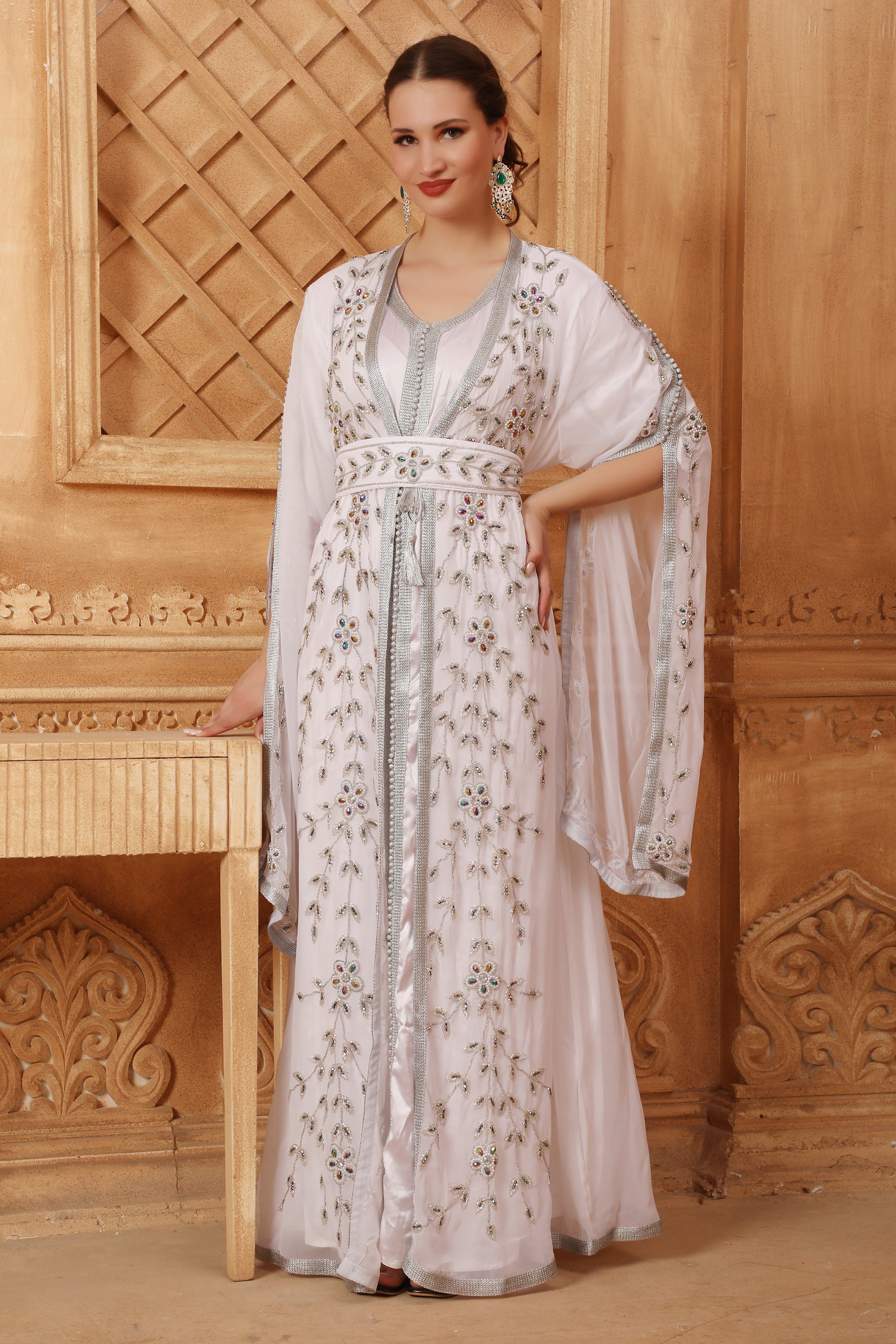 A woman wearing a white and silver Moroccan caftan with floral patterns and long sleeves, standing in a room with wooden decor.