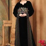 A woman standing in a beautifully embroidered black velvet gown with long straight sleeves, posing in a room with elegant decor.