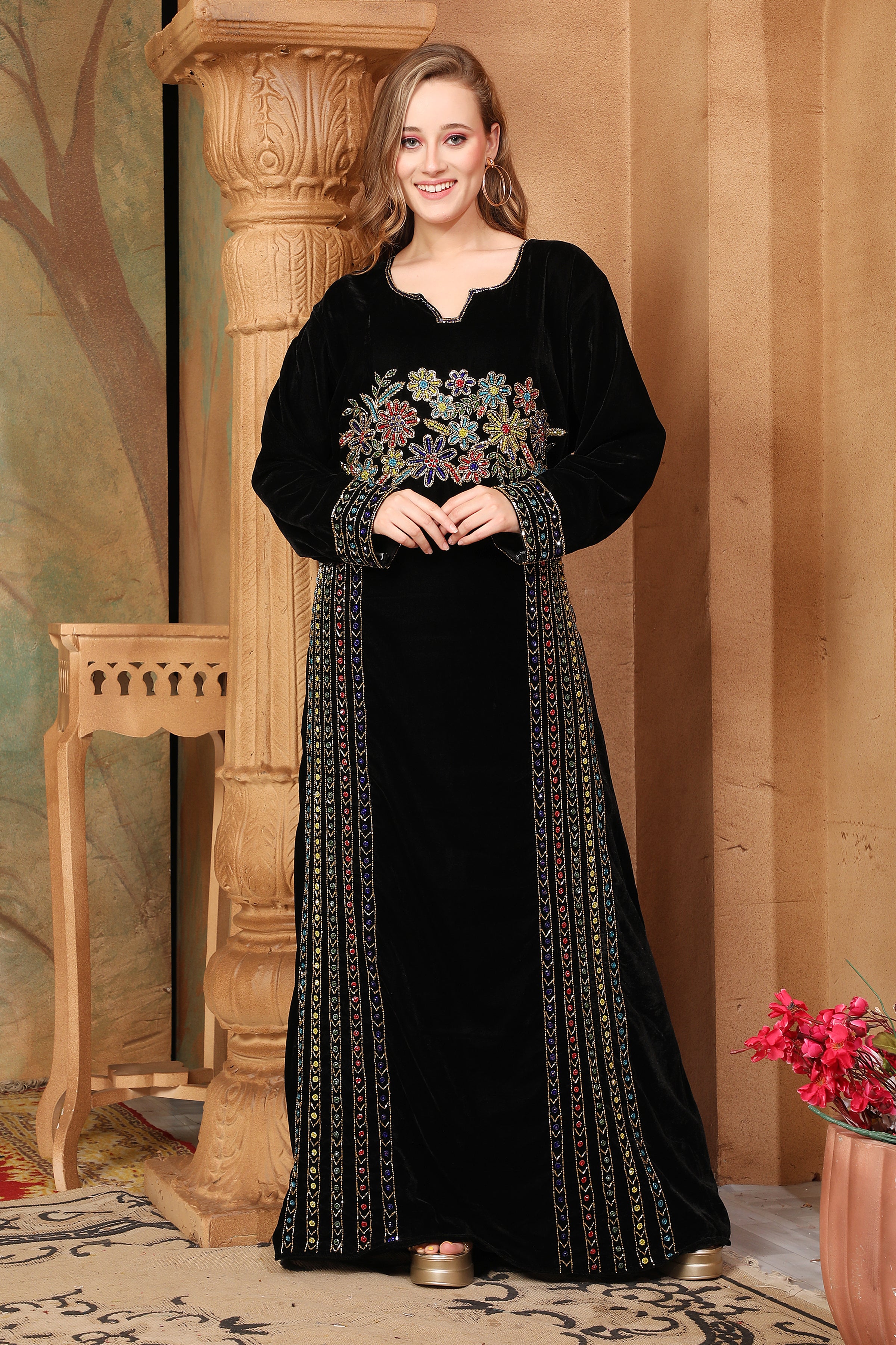 A woman standing in a beautifully embroidered black velvet gown with long straight sleeves, posing in a room with elegant decor.