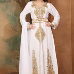 A woman standing in a beautifully embellished white Moroccan caftan with golden beads, posing against a background with trees.