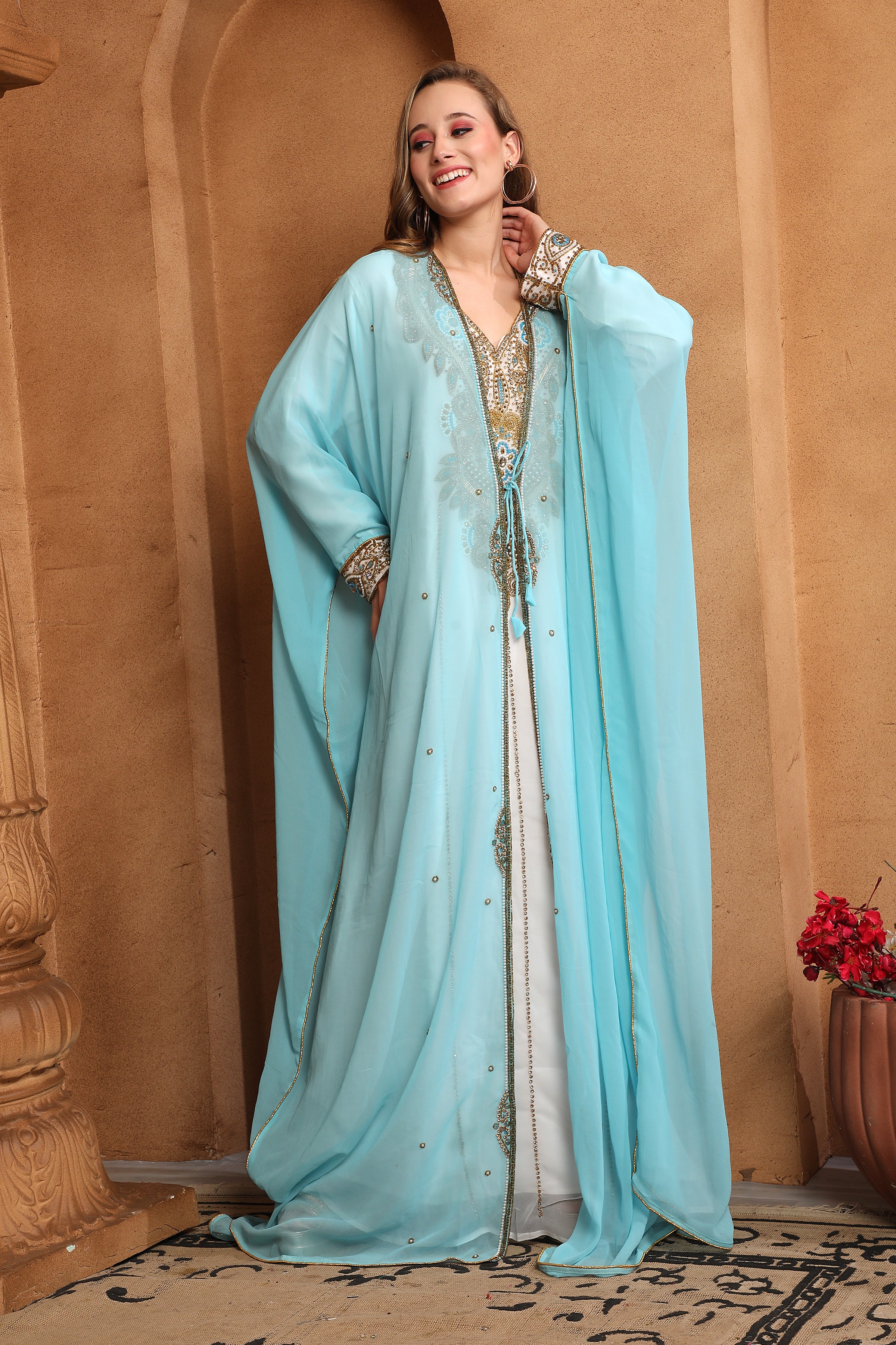 A woman wearing a blue and white traditional caftan with embellished golden beads, posing in a room with warm lighting.