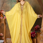 A woman standing in a beautiful yellow caftan with embellished golden beads, posing in a room with floral decorations and a wooden structure in the background.