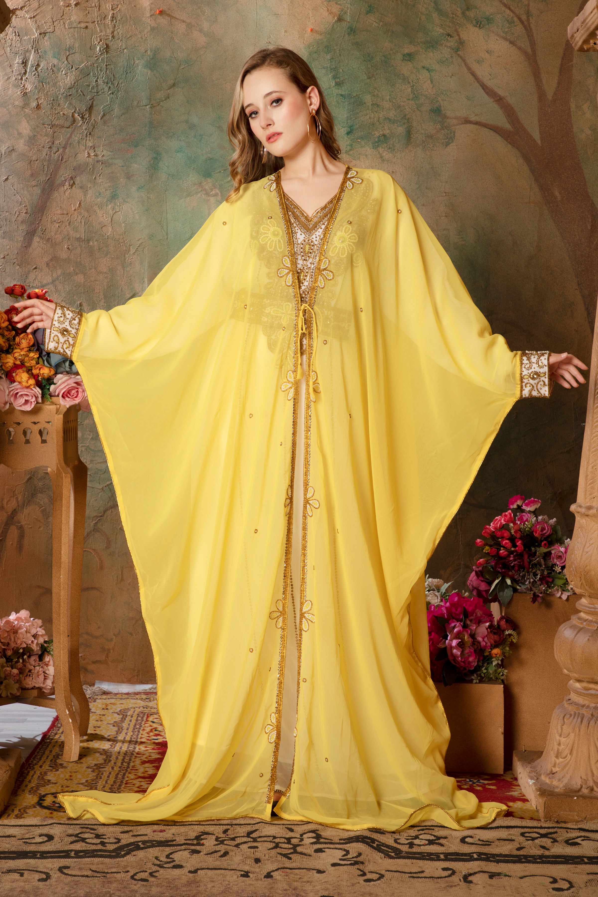 A woman standing in a beautiful yellow caftan with embellished golden beads, posing in a room with floral decorations and a wooden structure in the background.