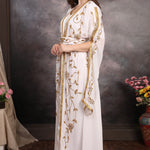 Woman in a white dress with gold embroidery standing against a textured wall.